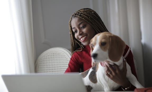 A happy woman with braided hair holding a dog while using a laptop indoors.